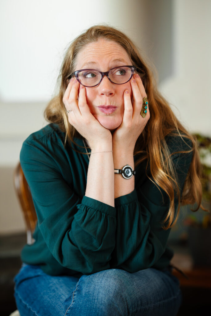 Ondine Delavelle in dunkelgrüner Bluse und Jeans sitzt auf einem Stuhl, stützt den Kopf in die Hände und schaut mit leicht verschmitztem Lächeln nach oben-links; sichtbar sind lange Haare, eine Uhr und ein Ring mit türkisfarbenen Steinen.