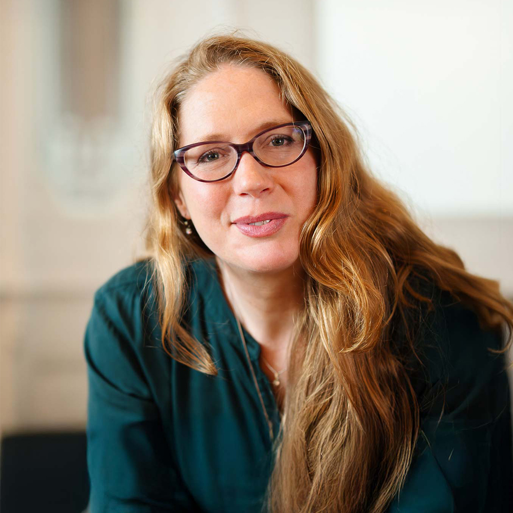 Willkommens-Portrait von Ondine Delavelle in dunkelgrüner Bluse in einem hellen Raum, mit sanftem Lächeln, leicht nach vorn gelehnt, als würde sie einem Gegenüber aufmerksam zuhören