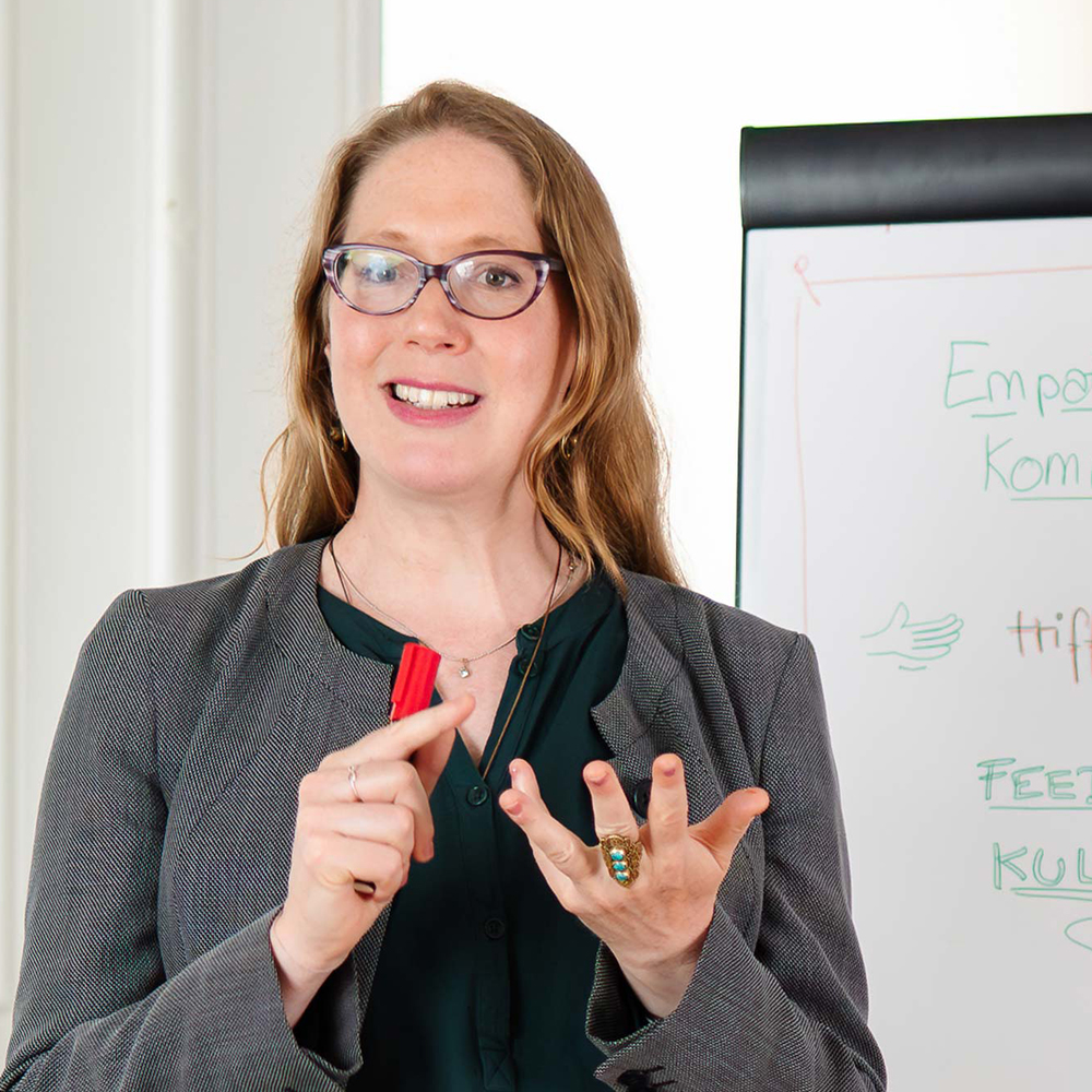 Ondine Delavelle, mit grauem Blazer und dunkelgrüner Bluse, steht vor einem Flipchart und erklärt Inhalte zu empathischer Kommunikation, hält einen roten Stift in der Hand und zählt mit den Händen auf.