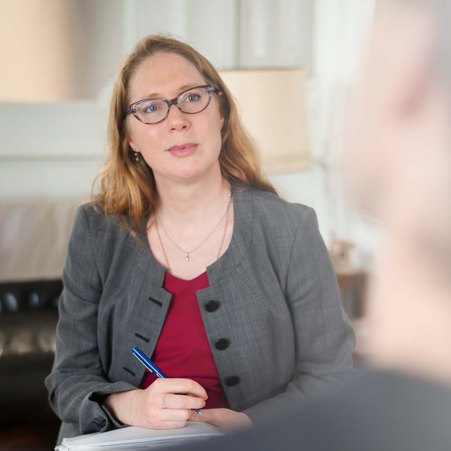 Ondine Delavelle sitzt in einem grauen Blazer und bordeauxroten Oberteil mit Notizbuch und Stift vor sich und hört einer gegenüber sitzenden Person aufmerksam zu; im Hintergrund sind Sofa und Lampe zu erkennen.