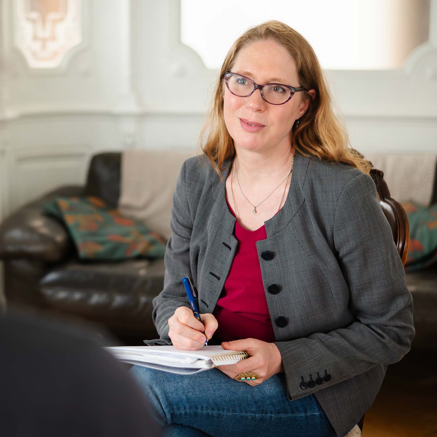 Ondine Delavelle sitzt in einem grauen Blazer, pinkem Oberteil und Jeans auf einem Stuhl, hält ein Notizbuch auf dem Schoß und schreibt mit einem Stift, während sie einer Person gegenüber aufmerksam zuhört; im Hintergrund steht ein dunkles Sofa mit Kissen.