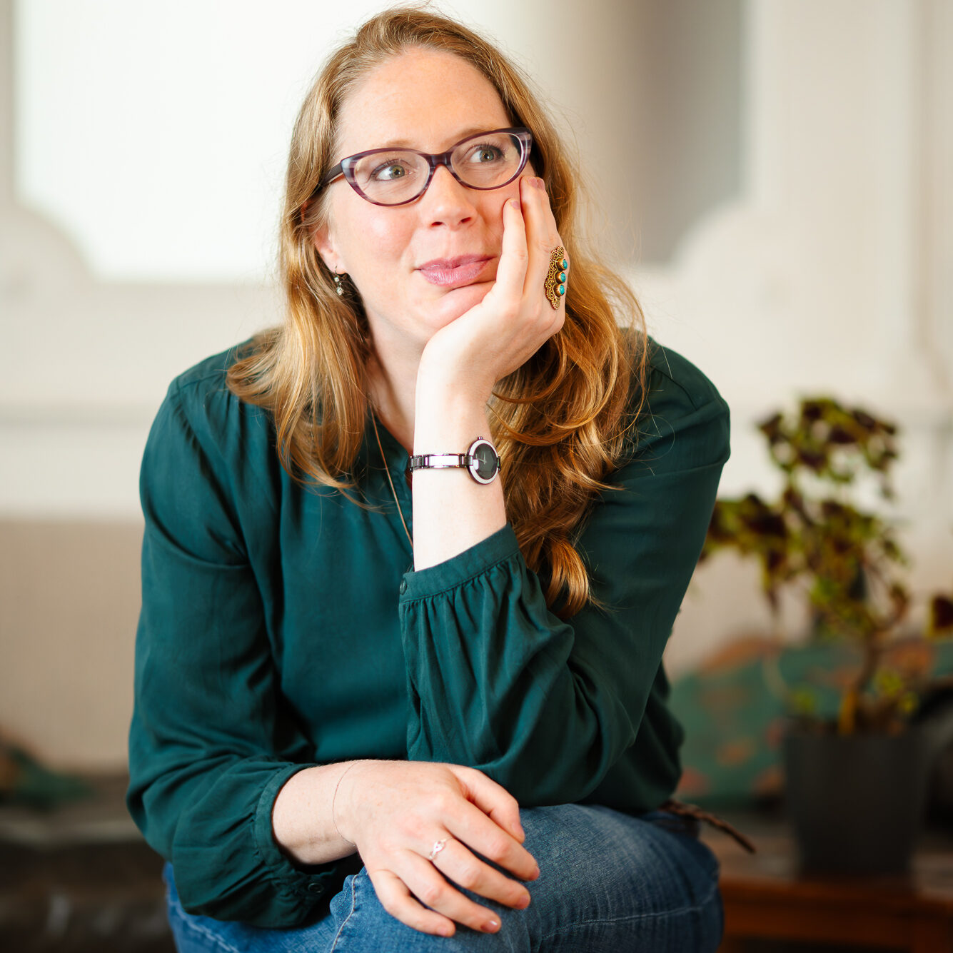 Trainerin Ondine Delavelle mit offenen Haaren in einer dunkelgrünen Bluse und Jeans sitzt mit überschlagenen Beinen auf einem Stuhl, stützt den Kopf auf die linke Hand, im Hintergrund sind Sofa und Zimmerpflanze zu sehen.