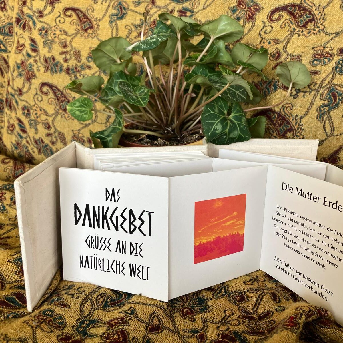 Auf einem ocker gemusterten Sofa steht ein aufgeklapptes, handgebundenes Leporello-Büchlein mit der Aufschrift ‚Das Dankgebet – Grüße an die natürliche Welt‘ neben einer grünen Zimmerpflanze; rechts ist eine Seite mit dem Text ‚Die Mutter Erde‘ zu sehen.